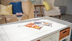 Ein weißer Couchtisch mit zwei Weingläsern und Zeitschriften in einem Wohnzimmer mit beiger Couch, blauen und gelben Kissen und einem weißen Sessel.