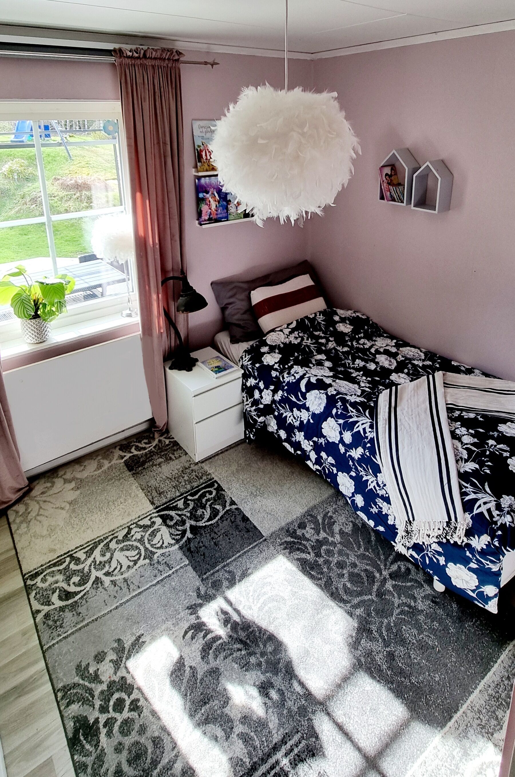 Une chambre à coucher comprenant un lit, une table de chevet, une fenêtre avec des rideaux, un luminaire suspendu et une étagère fixée au mur avec des livres.