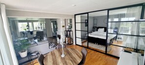 Quarto de hotel moderno com uma grande porta de vidro deslizante que dá para uma varanda, uma área de jantar separada com uma mesa redonda de madeira, e um espaço de estar acolhedor com um sofá e uma pequena mesa lateral.