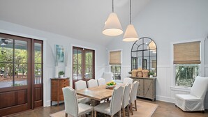 A dining area with a wooden table, white chairs, and pendant lights. There is a view of the outdoors through large windows.