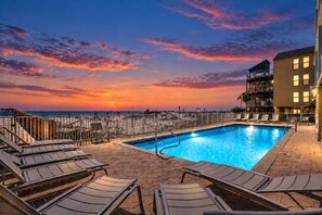 Une piscine avec des chaises longues, une terrasse en bois et une vue sur l’océan au coucher du soleil.