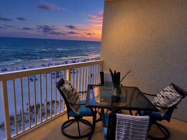 A balcony with a glass table and chairs, overlooking a beach with people and umbrellas.