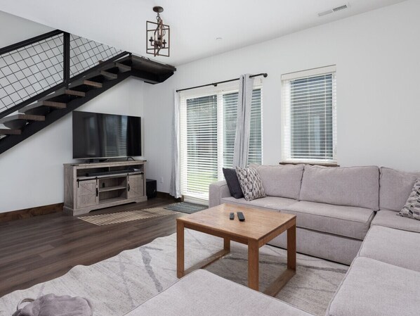 A modern living room with a staircase, a flat-screen TV, a grey sofa, and a coffee table.