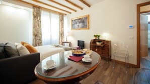 A hotel room with a bed, a sofa, a glass coffee table, a TV, a wooden dresser, and a clear plastic chair. - Casa Sorelle Flo - Flo Apartments (Firenze)
