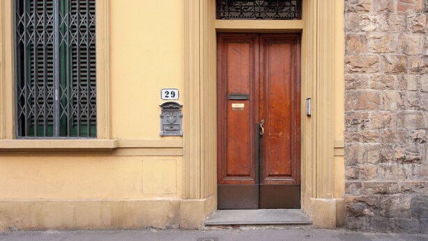 A building with a yellow facade, a stone wall, and a wooden double door with a metal handle. - Cate Apartment - Flo Apartments (Firenze)