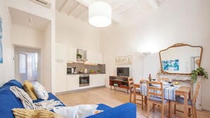 A modern living room with a blue sofa, wooden dining table, and chairs. There is a kitchen area with white cabinets and a large mirror.