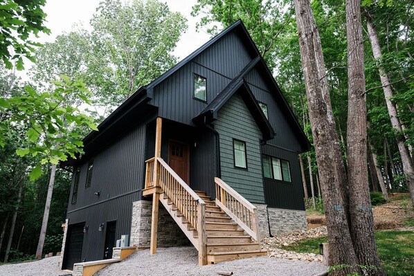 A modern two-story cabin with a wooden staircase, stone foundation, and a steeply pitched roof, surrounded by a forest.