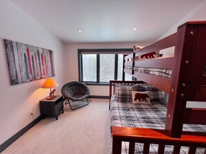 Un dormitorio con literas, una silla, una lámpara, una obra de arte en la pared y una ventana con vistas a árboles.