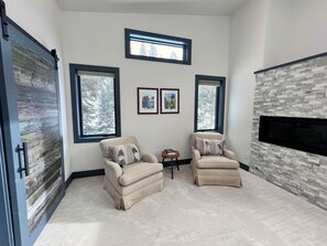 Una habitación con dos sillones color beige, una pequeña mesa, obras de arte enmarcadas en la pared y una chimenea con textura de piedra.