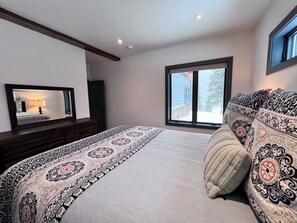 Un dormitorio con una cama grande, una mesita de noche, un espejo y una ventana con vistas a un edificio y árboles.
