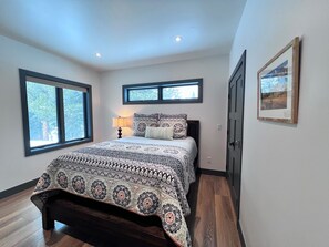 Un dormitorio con una cama grande, dos ventanas, una mesita de noche con una lámpara y un cuadro enmarcado en la pared.