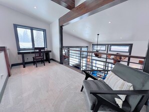 Una habitación amplia con techo alto, un escritorio con silla, un sofá y vistas al exterior a través de un gran ventanal.