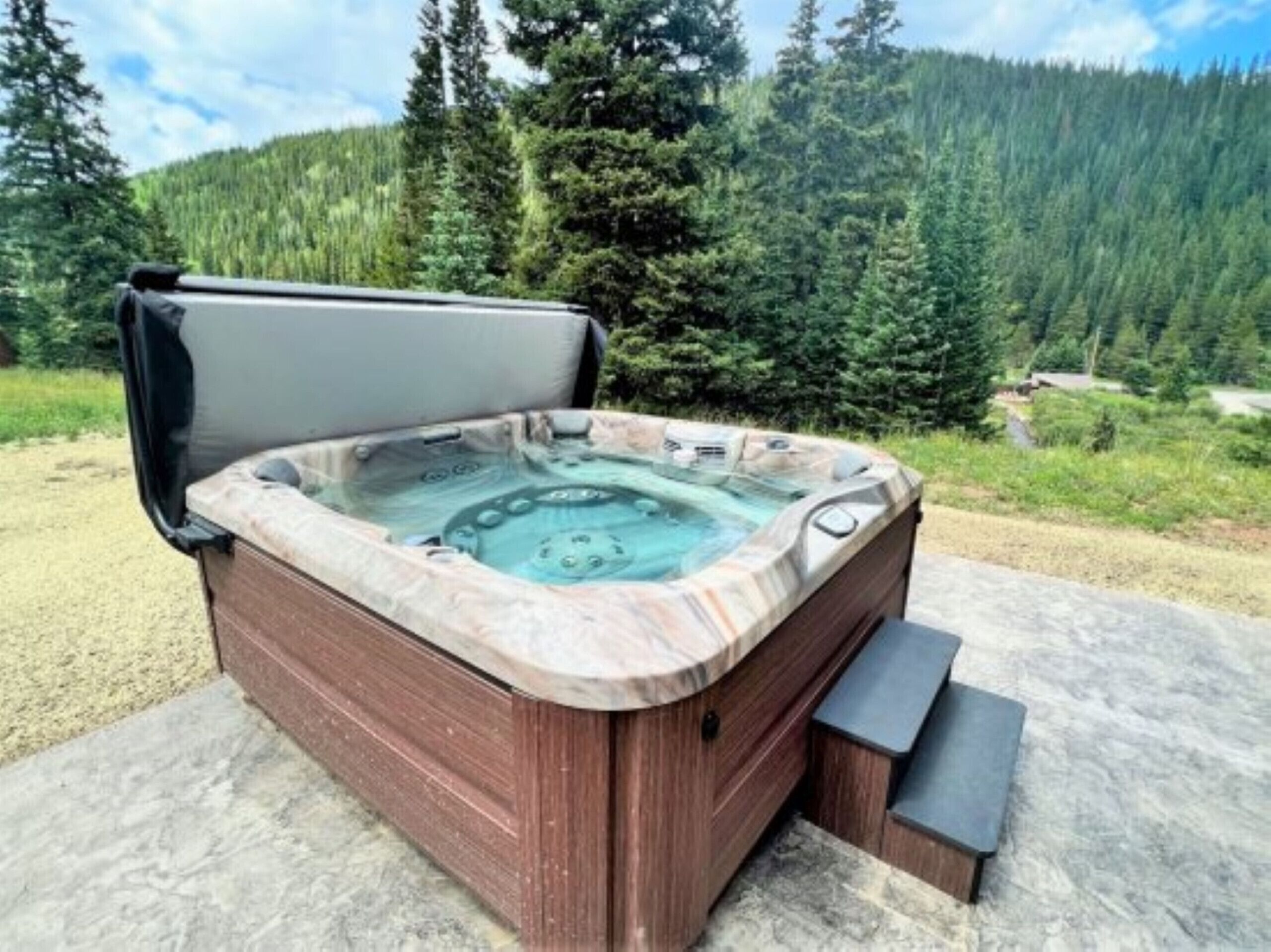 Un bain à remous doté d’une structure extérieure en bois et d’un intérieur imitant la pierre, installé sur une surface en béton, avec une montagne boisée en toile de fond.