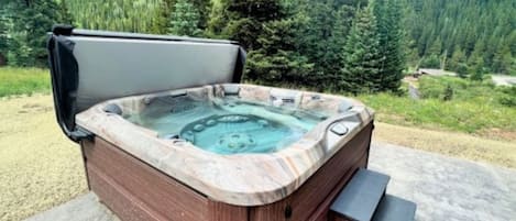 A hot tub with a wooden exterior and a stone-like interior, set on a concrete surface with a forested mountain backdrop.