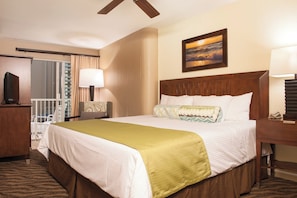A hotel room with a large bed, a wooden headboard, a painting of a sunset, a desk with a lamp, and a view of the city through the window.