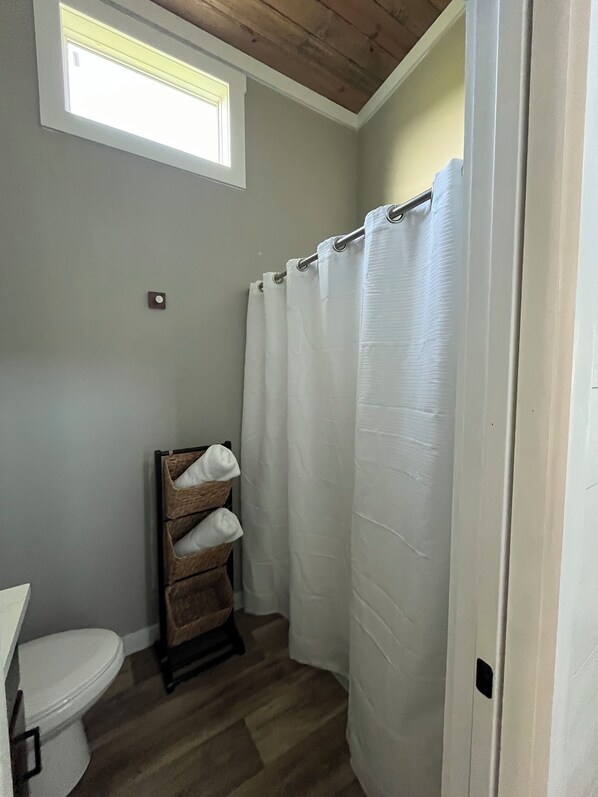 A bathroom with a wooden floor, a white toilet, a towel rack with folded towels, a shower curtain, and a small window above the toilet. - Circle T Mini Farms (Strawberry Plains)