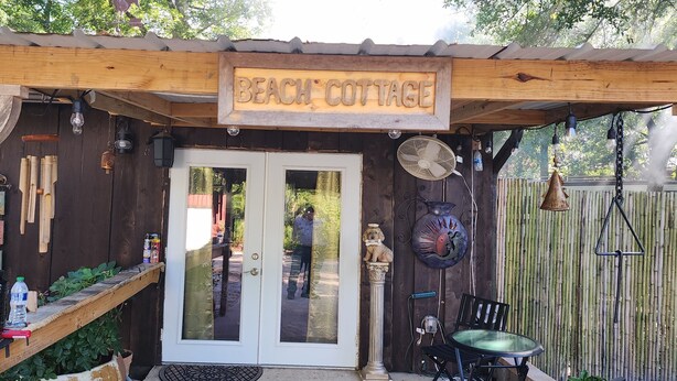 Una estructura de madera con un letrero que dice "Beach Cottage", una puerta de cristal, un ventilador y una pequeña mesa con una silla.