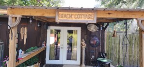 A wooden structure labeled "Beach Cottage" with a glass door, a fan, and a small table with a chair.