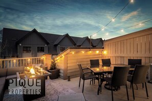 Une terrasse avec une table à plateau de verre, des chaises, un foyer et des guirlandes lumineuses.