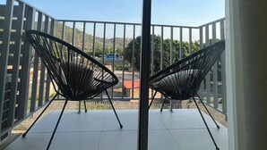 A balcony with two black chairs, a white table, and a view of a parking lot and hills.