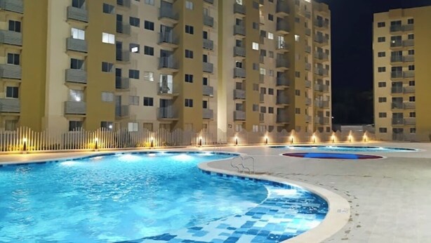 A swimming pool with tiled flooring, surrounded by a fence, and adjacent to a multi-story building with balconies.