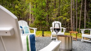 Un rincón exterior acogedor con sillas Adirondack, un hogar de fuego metálico y una copa de vino tinto.