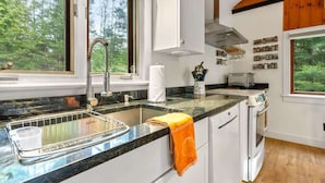 Cocina con encimera de granito, pileta de acero inoxidable y muebles blancos. Se ve una ventana con vista a árboles, y una toalla colgada en la puerta del horno.
