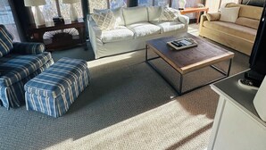 Una sala de estar acogedora con un sofá blanco, un reposapiés a rayas y una mesa de centro de madera. Hay dos lámparas sobre mesas auxiliares, y una mesita con una lámpara y un teléfono.