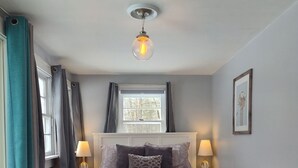 Un dormitorio con cama, lámparas de noche, una ventana con cortinas, una lámpara de techo e imagen enmarcada en la pared.