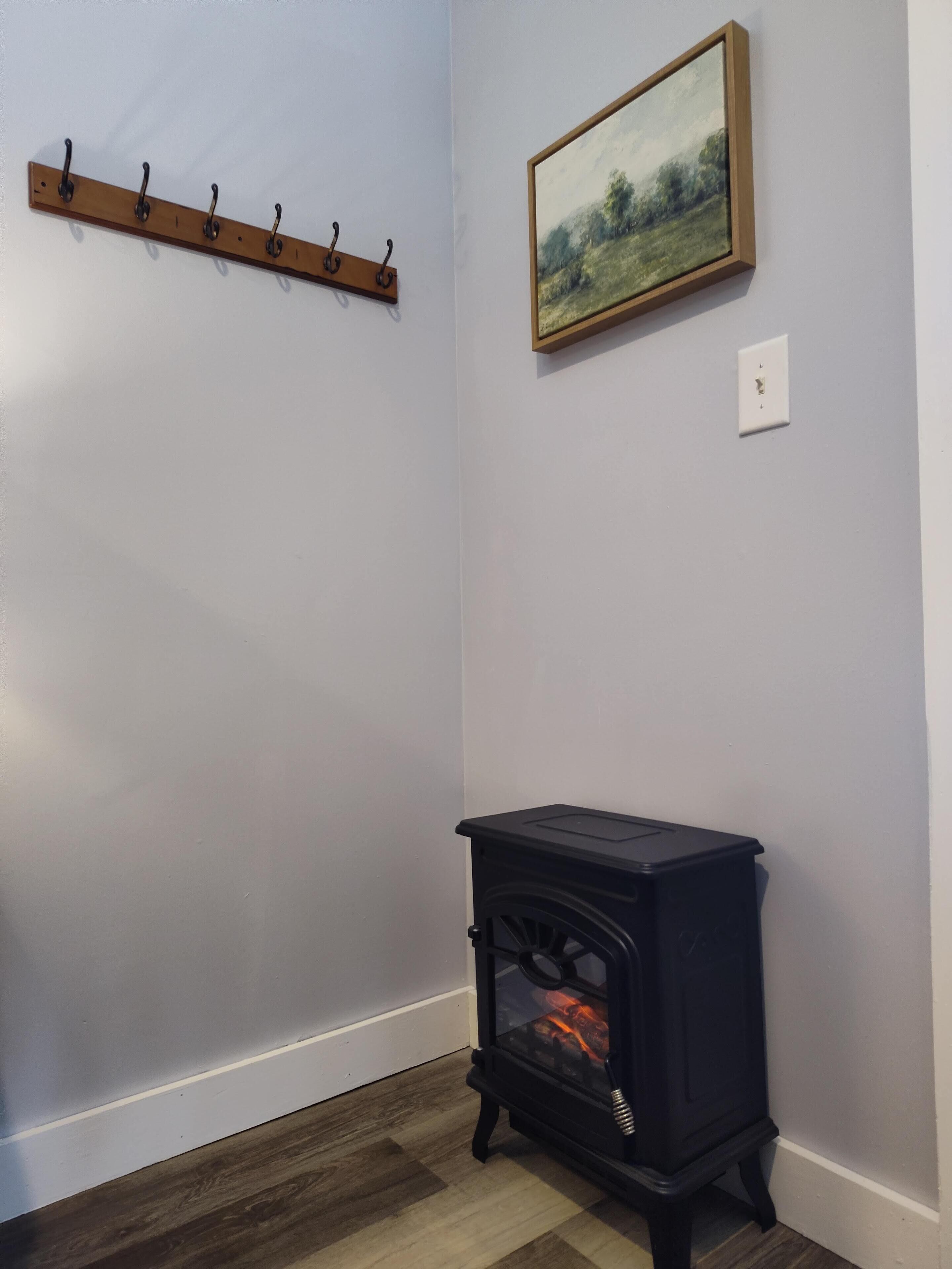 A corner room with a black wood-burning stove, a coat rack, and a framed landscape painting on the wall.
