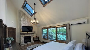 A bedroom with a stone fireplace, a TV mounted above it, a large bed with white bedding, and skylights. - Lakefront Retreat with Private Dock and Hot Tub (Lackawaxen)