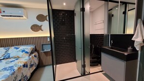 Quarto de hotel moderno com cama, escrivaninha pequena, banheiro com vaso sanitário e pia, e um espelho grande.
