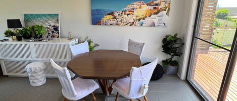 A dining area with a round wooden table and white upholstered chairs. There is a painting of a coastal village on the wall.