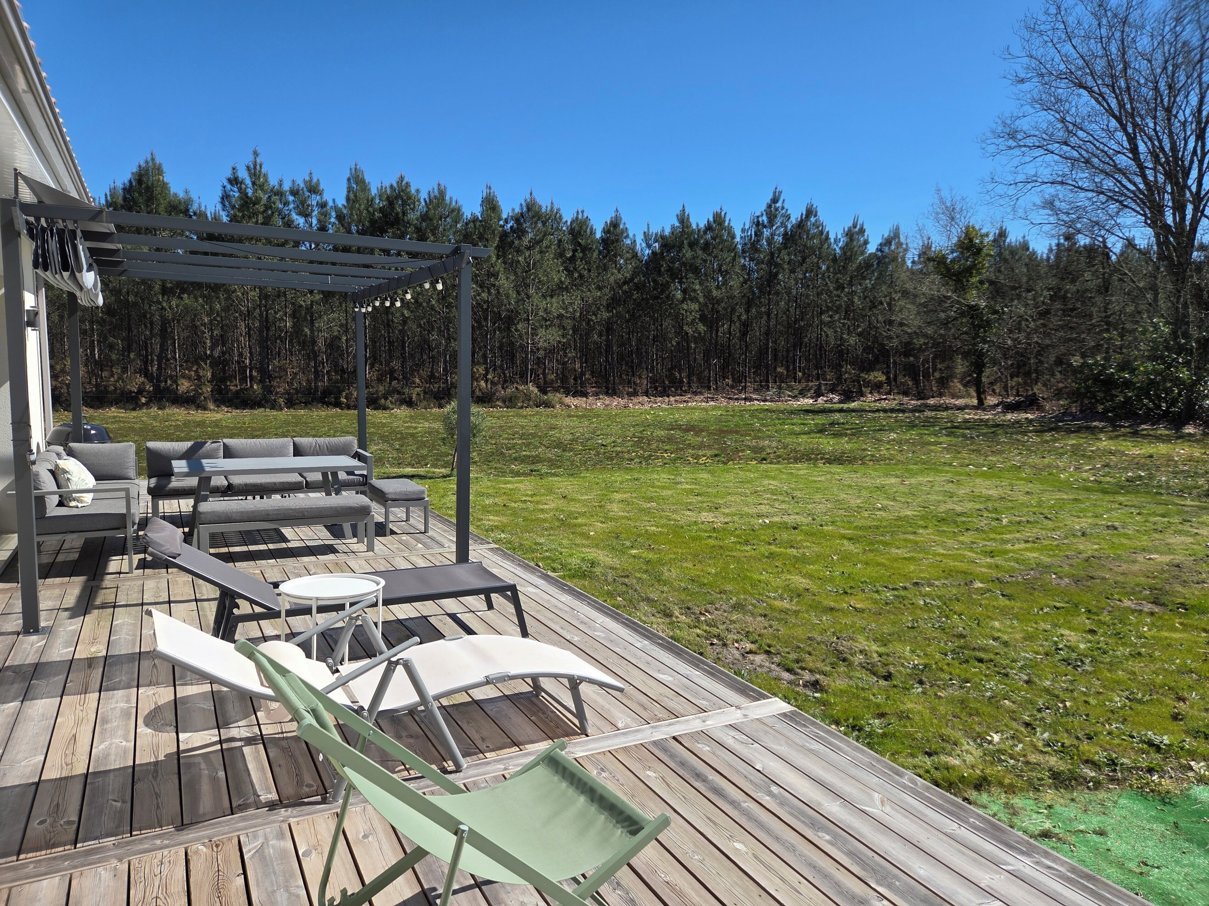 Eine Holzterrasse mit Gartenmöbeln, darunter eine Sitzecke und Stühle, unter einer Pergola.