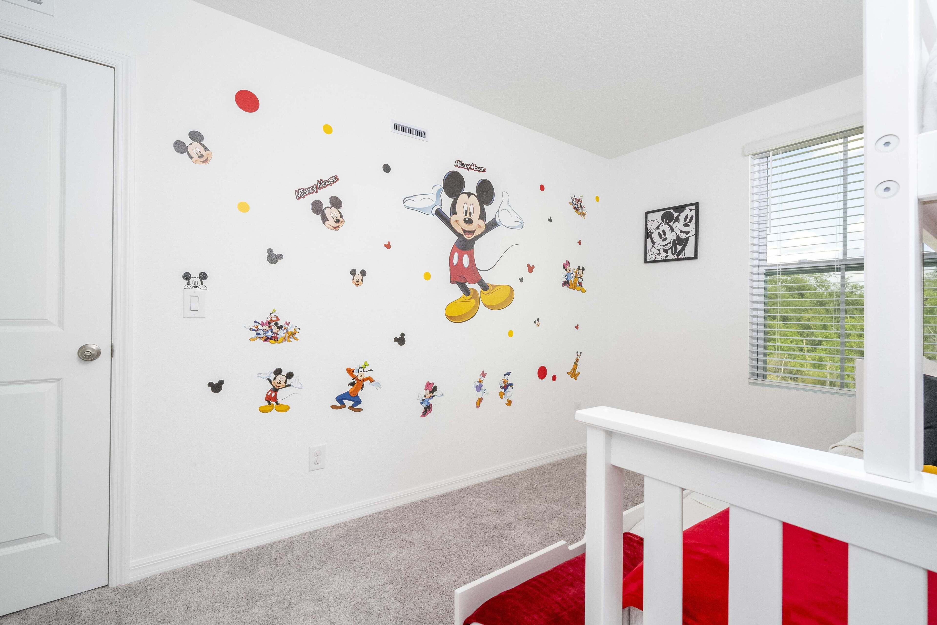 A room with white walls decorated with Mickey Mouse decals, a white door, a window with blinds, and a red and white crib.
