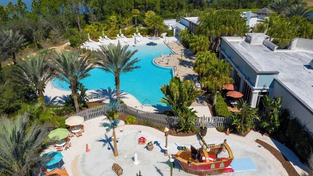 An aerial view of a resort with a large swimming pool, palm trees, and a play area with a pirate ship. - Cozy 3Bd W/ Free Waterpark @ Enclaves At Festival 713 (Davenport)