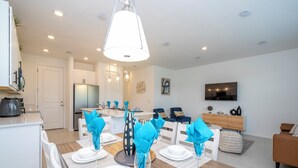 A modern dining area with a white table set for six, a kitchen in the background, and a living area with a sofa and TV. - Cozy 3Bd W/ Free Waterpark @ Enclaves At Festival 713 (Davenport)
