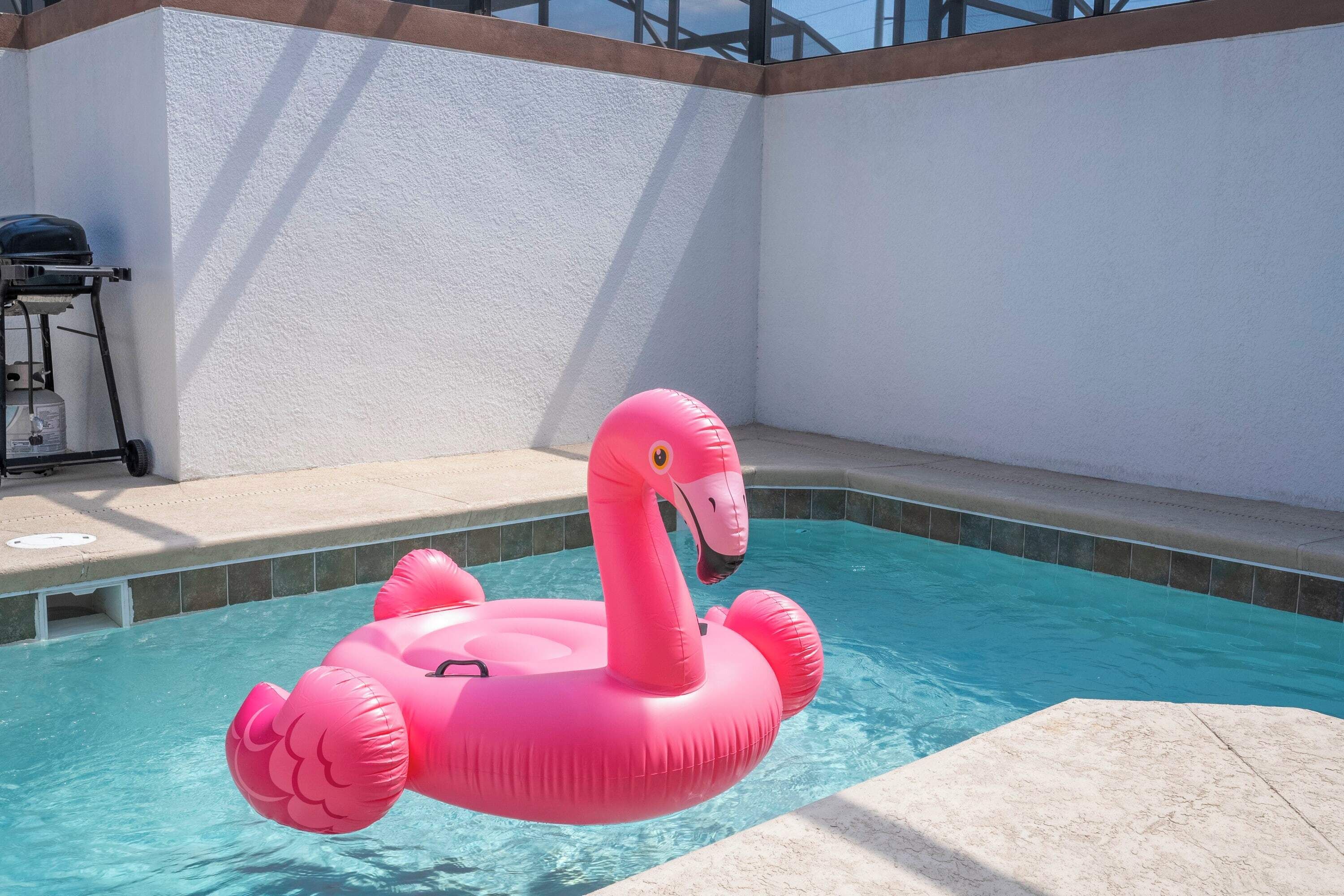 A pink flamingo float in a pool with a white wall and a grill in the background.