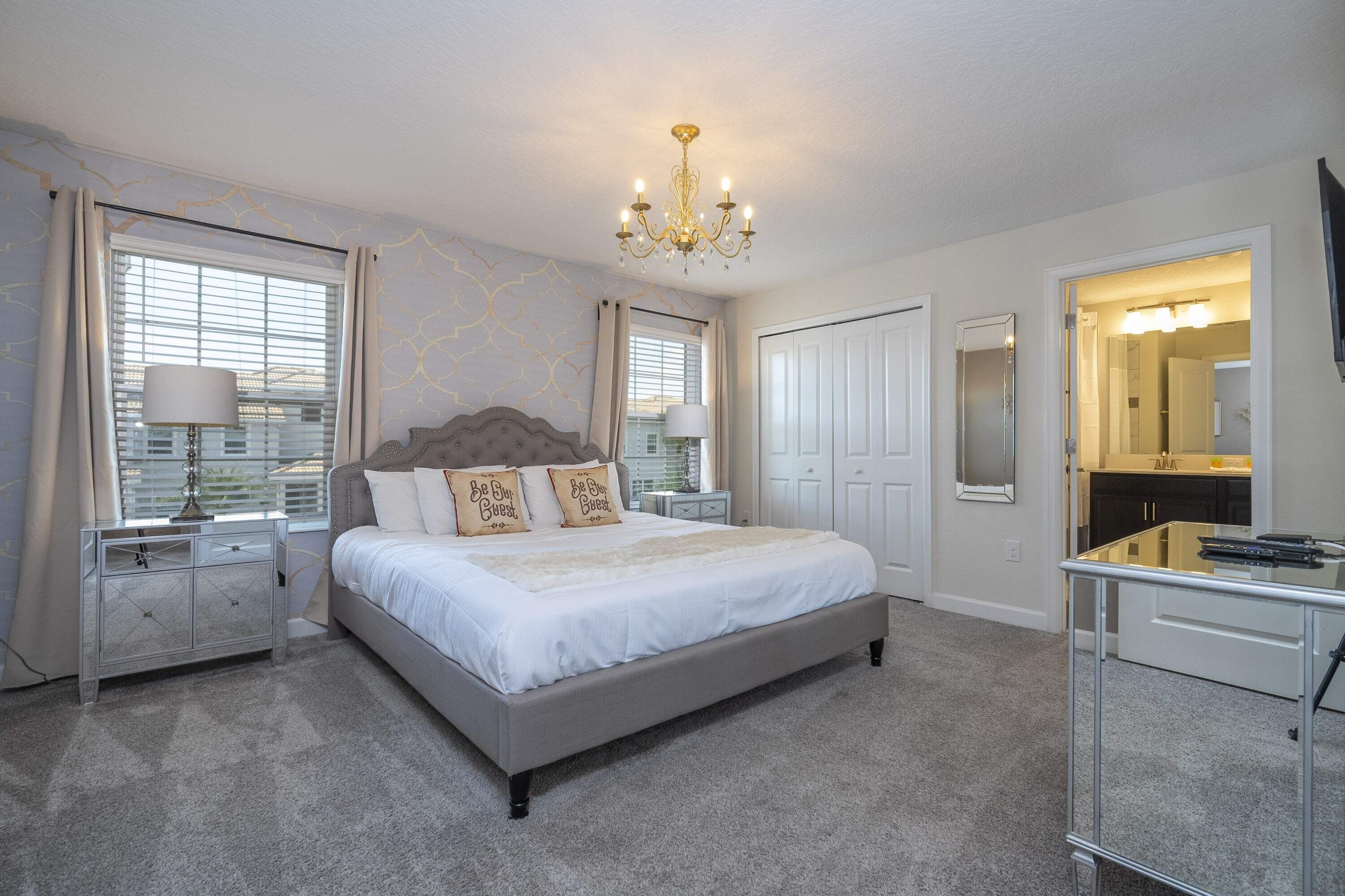 A bedroom with a large bed, bedside tables, a dresser, a chandelier, and a bathroom visible through an open door.