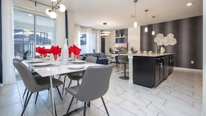 A modern kitchen with a central island, dining area with chairs and a table, and a living area with a sofa and a TV. - Gorgeous 4Bd W/ Pool & Waterpark @ Storey Lake 4836M (Kissimmee)