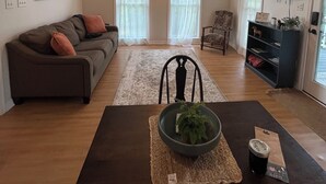 En stue med et mørkebrunt sofabord, en grå sofa med puder, en sort lænestol, et fjernsyn på væggen og en loftventilator.