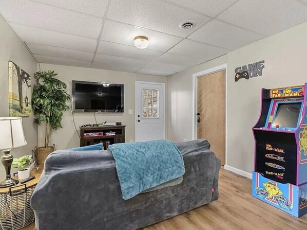 Un salón con un sofá gris, un televisor, una máquina arcade de Pac-Man y una puerta.