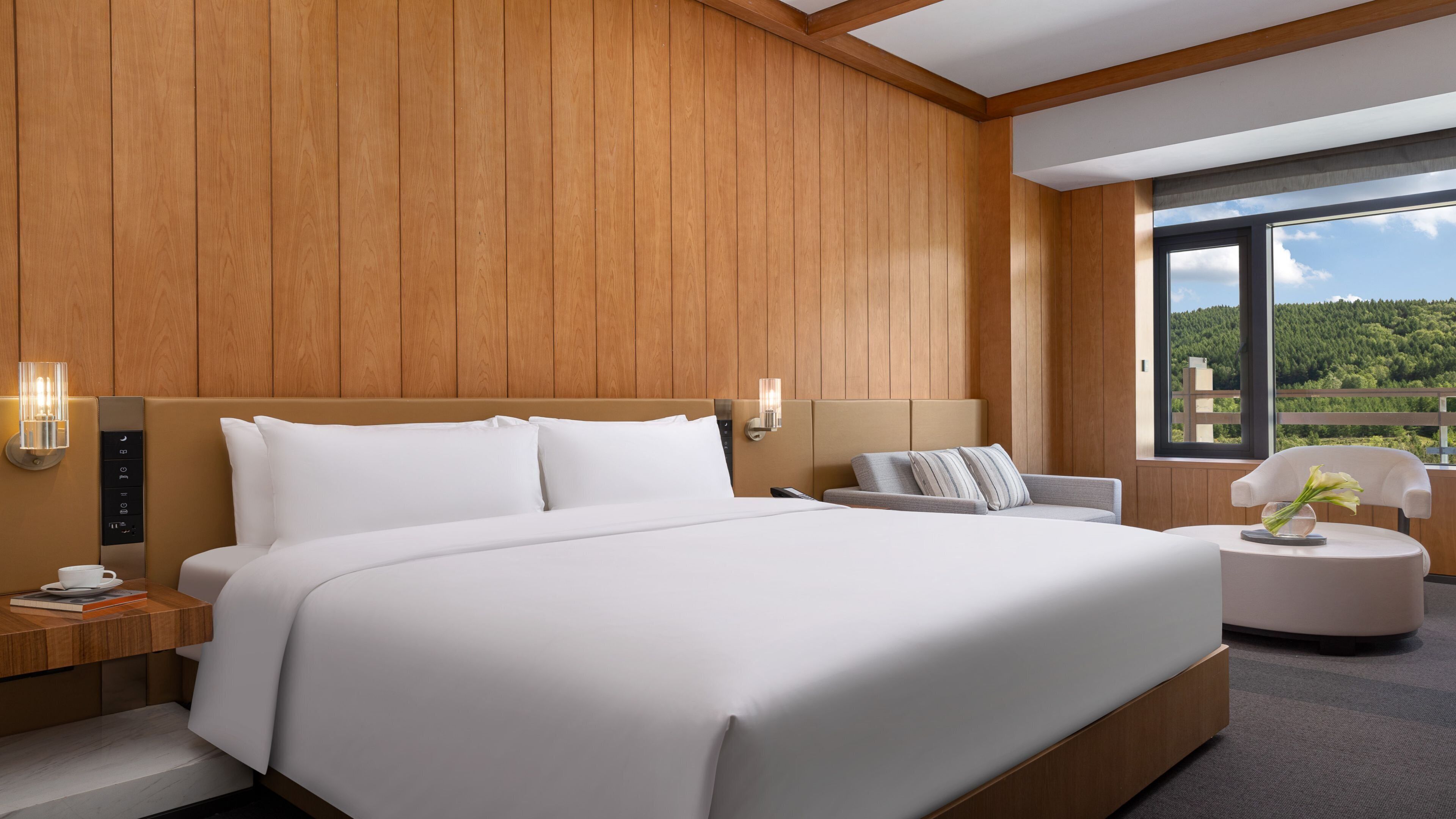 A modern hotel room with a large bed, bedside controls, a seating area with a chair and table, and a large window offering a view of a forested landscape.