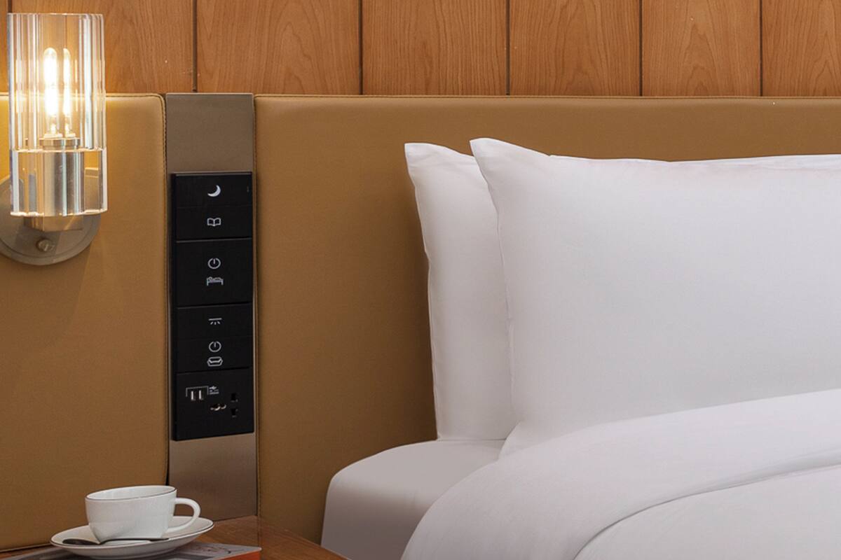 Une chambre d’hôtel avec un lit, une table de chevet sur laquelle se trouvent une tasse et une soucoupe, et un panneau de commande fixé au mur.
