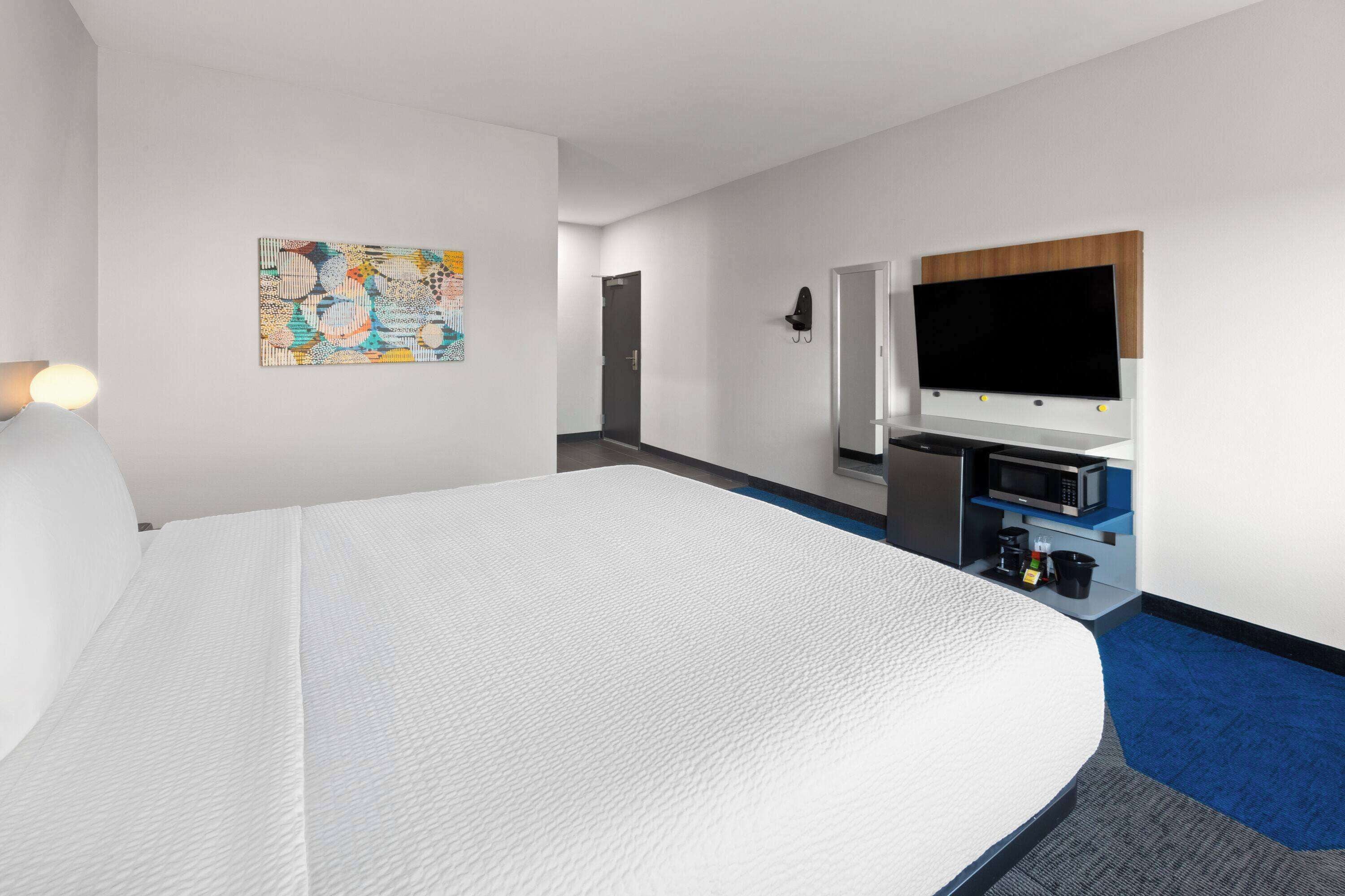A hotel room with a large bed, a flat-screen TV mounted on the wall, a microwave, and a small kitchenette area.