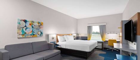 A hotel room with a bed, a sofa, a desk, a TV, and a painting on the wall.