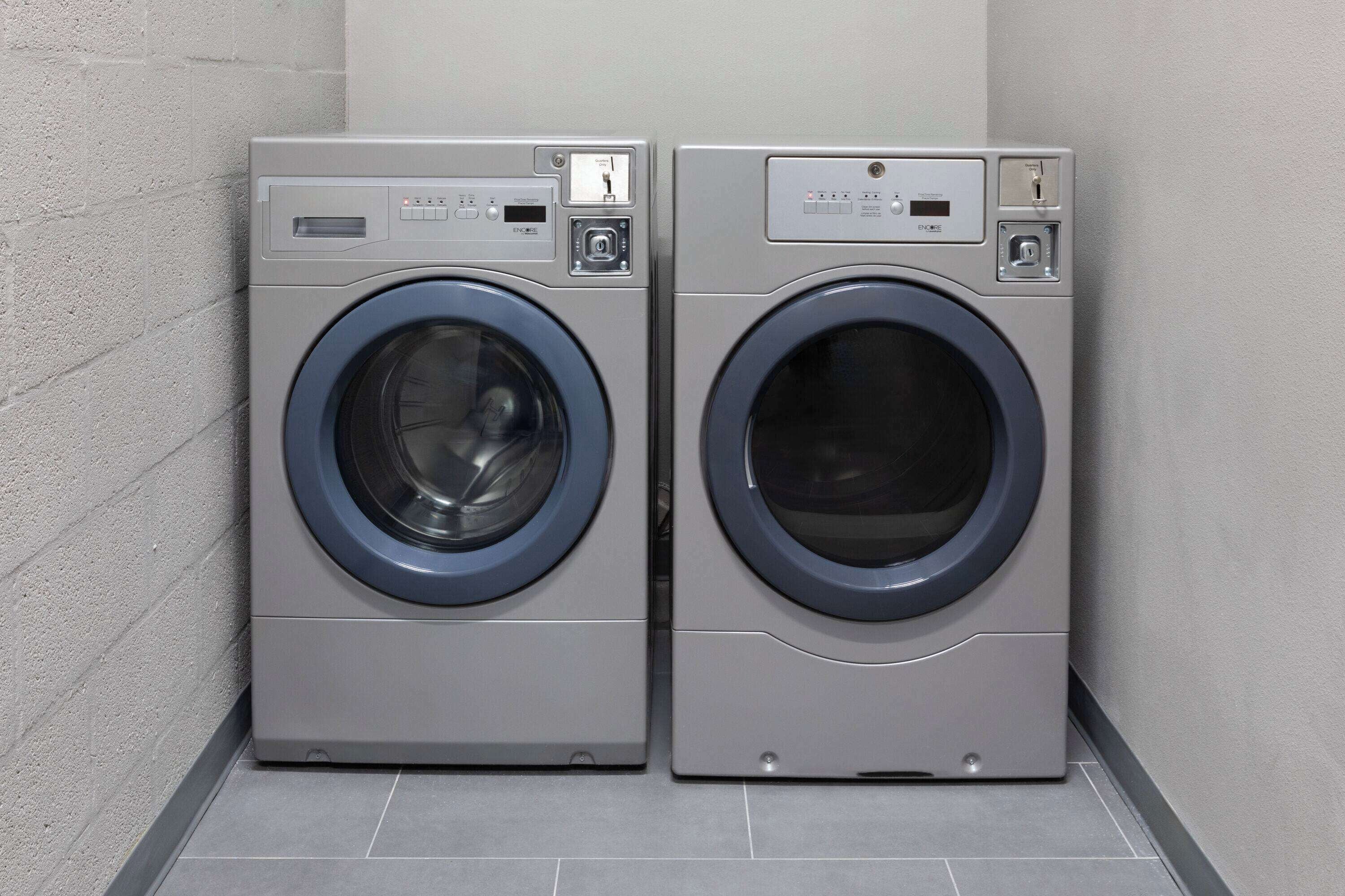 Two front-loading washing machines in a corner of a room with tiled flooring and a plain wall.
