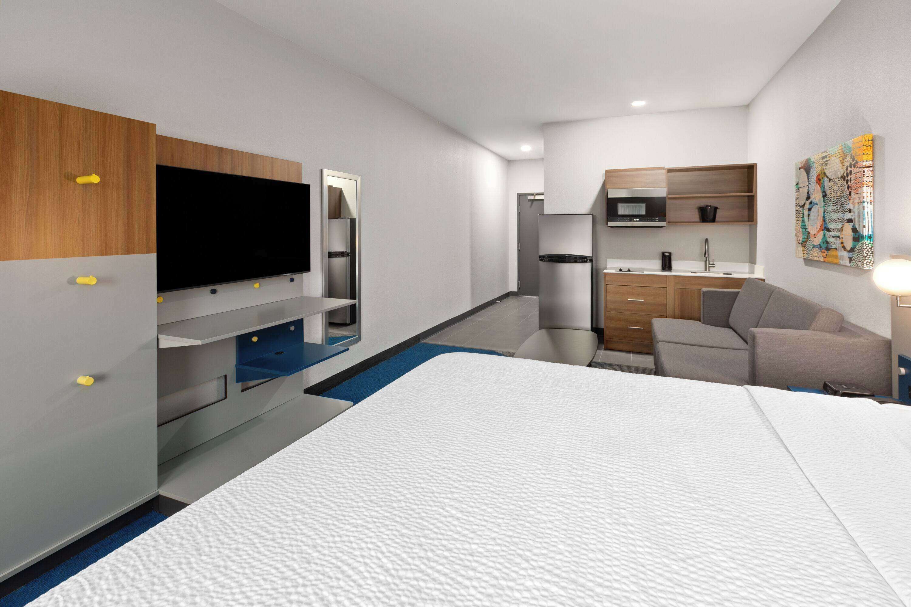 A modern hotel room with a bed, a TV, a kitchenette, and a seating area.
