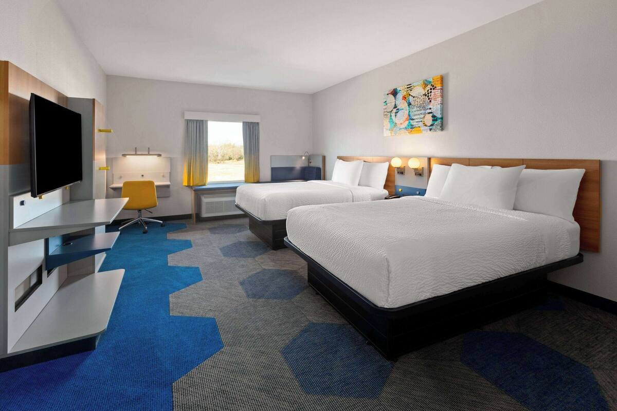 Une chambre d’hôtel avec deux lits, un bureau doté d’un téléviseur à écran plat, une chaise, une lampe de bureau et une fenêtre avec des rideaux.