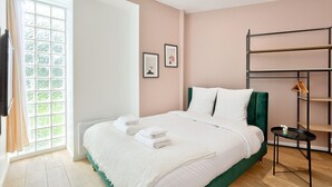 A neatly made bed with white linens and a green headboard, a window with a view of greenery, and a black metal shelf with hangers.